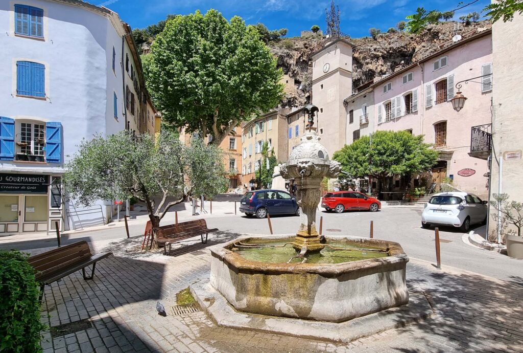 Een sfeervolle fontein in Cotignac, omringd door historische huizen, met de imposante rotswand op de achtergrond.